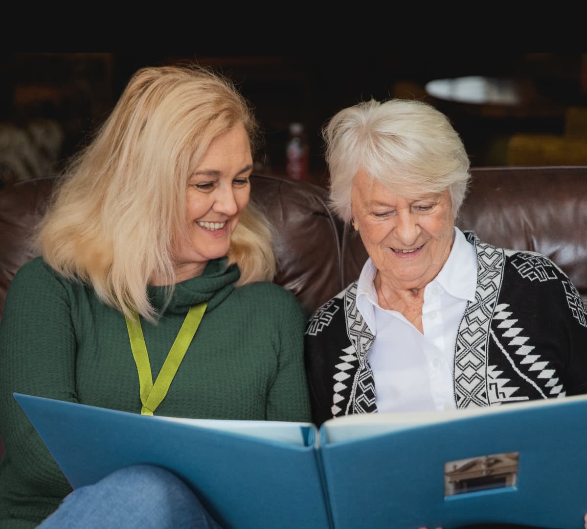 Two women happy and smiling while sitting on a couch and looking at a photo album inside the house