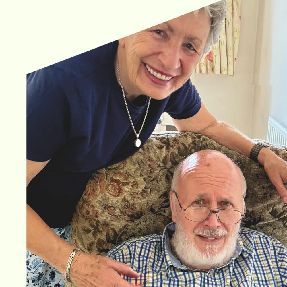 Smiling woman stands beside an older man seated on a couch; both appear happy in a cozy room. - Home Instead