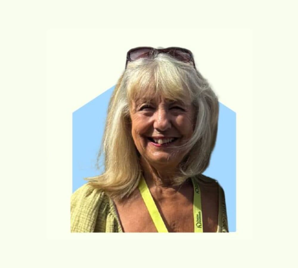 Smiling older woman with long blonde hair, sunglasses on head, and a yellow lanyard, against a light background. - Home Instead