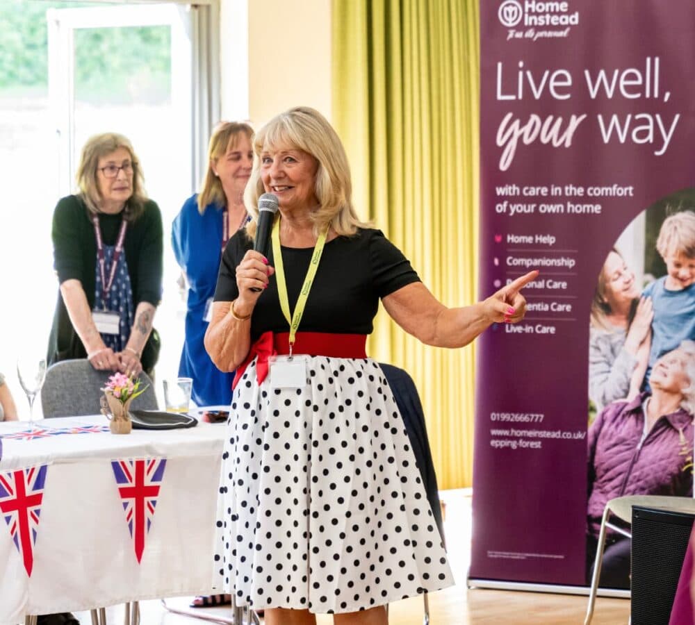 Smiling woman with a microphone speaks at an event, with people and a "Live well, your way" banner behind her. - Home Instead