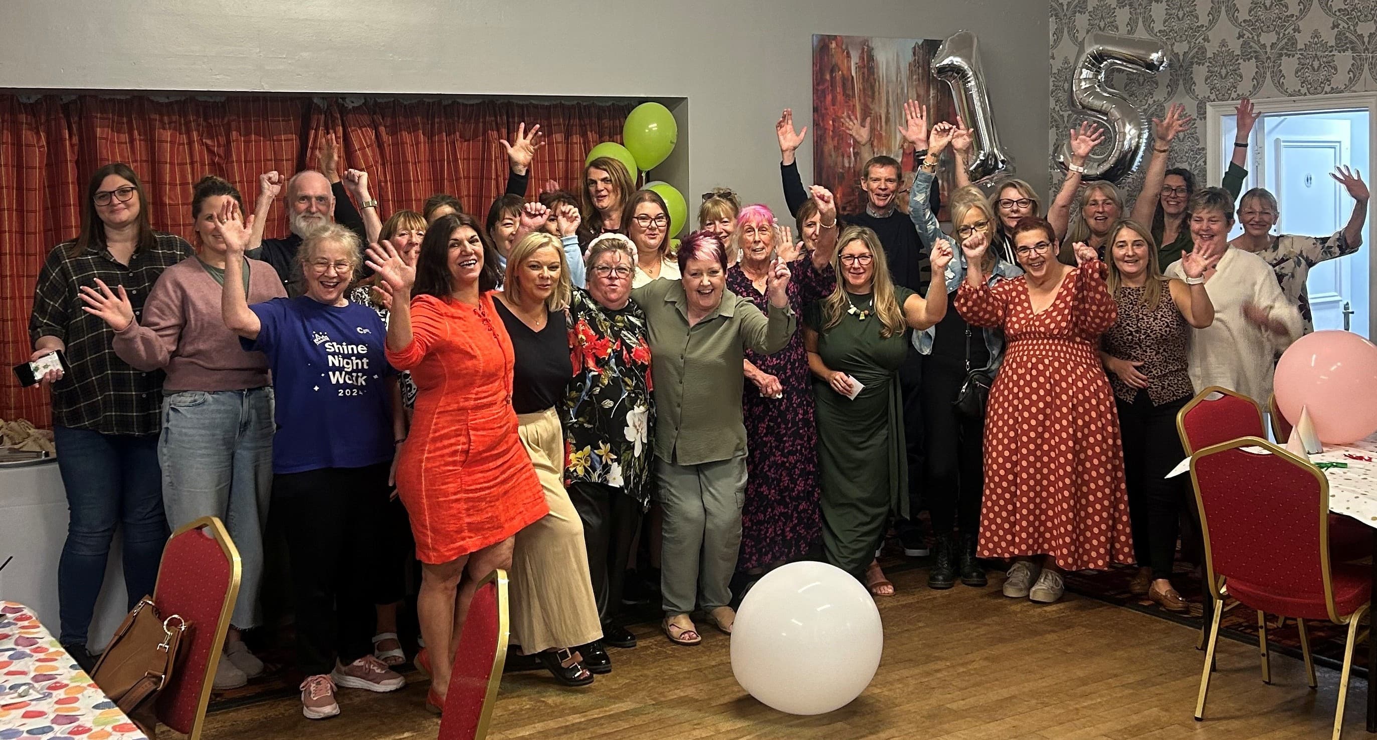 A large group of people smiling and waving at a party, with "15" balloons in the background. - Home Instead