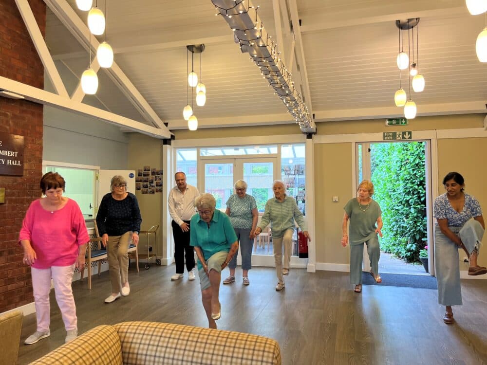 A group of seniors and an instructor practice gentle exercise together in a bright, airy room. - Home Instead