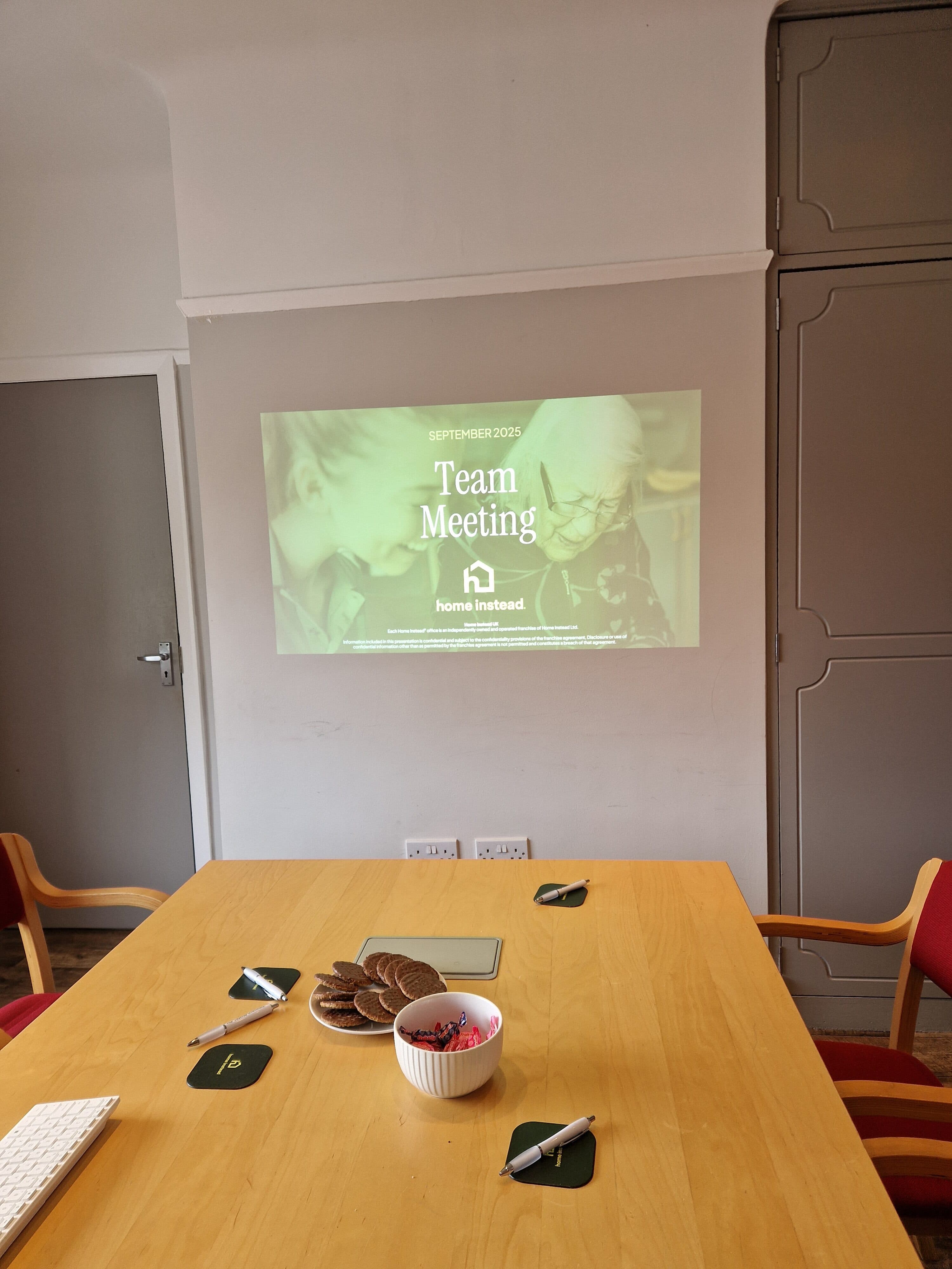 A projector displays "Team Meeting" on a wall above a table with snacks and stationery in a meeting room. - Home Instead