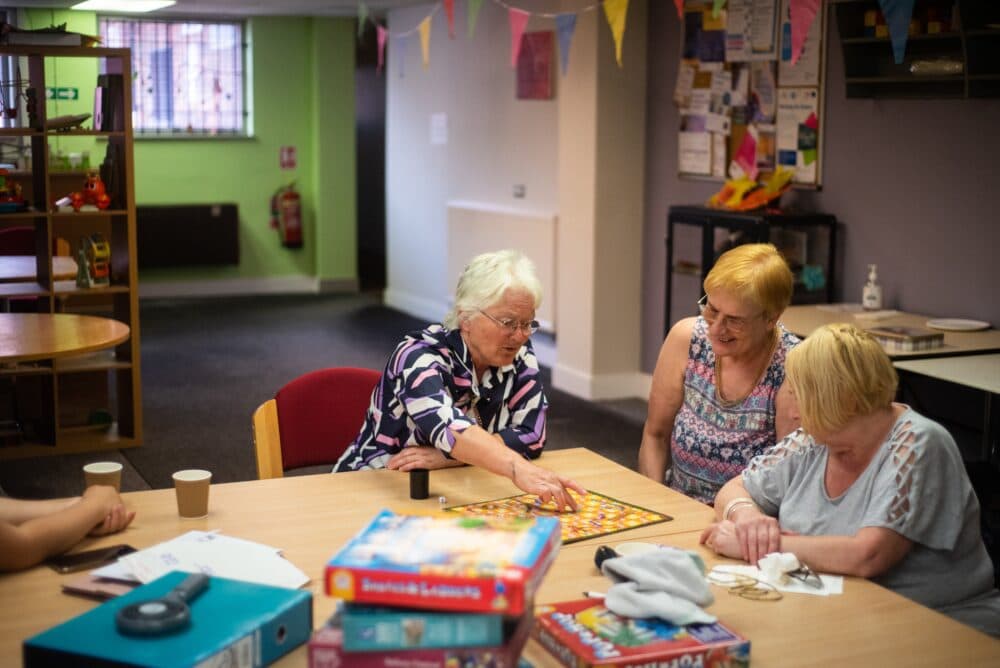 Three older women sit around a wooden table in what appears to be a community center or activity room, engaged in playing a board game together. The woman on the left wears glasses and a colorful striped top, while the woman in the middle has short blonde hair and wears a patterned sleeveless top. A third woman with blonde hair sits on the right in a light blue top. The table has coffee cups, game boxes, and what looks like a colorful board game spread out. The room has bright green and white walls, colorful bunting hanging from the ceiling, bulletin boards, and shelving with various items, creating a cheerful, social atmosphere typical of a senior center or community gathering space.