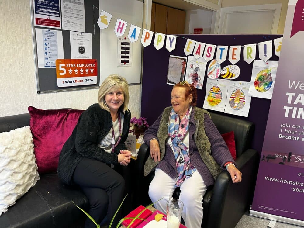 Two women sit and smile on a couch in a decorated room with an Easter banner and colorful drawings behind them. - Home Instead