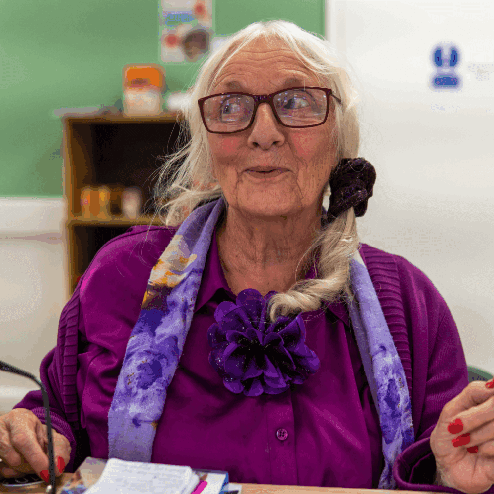 Elderly woman with long blonde hair and glasses wearing purple clothing sits at desk in classroom or office setting