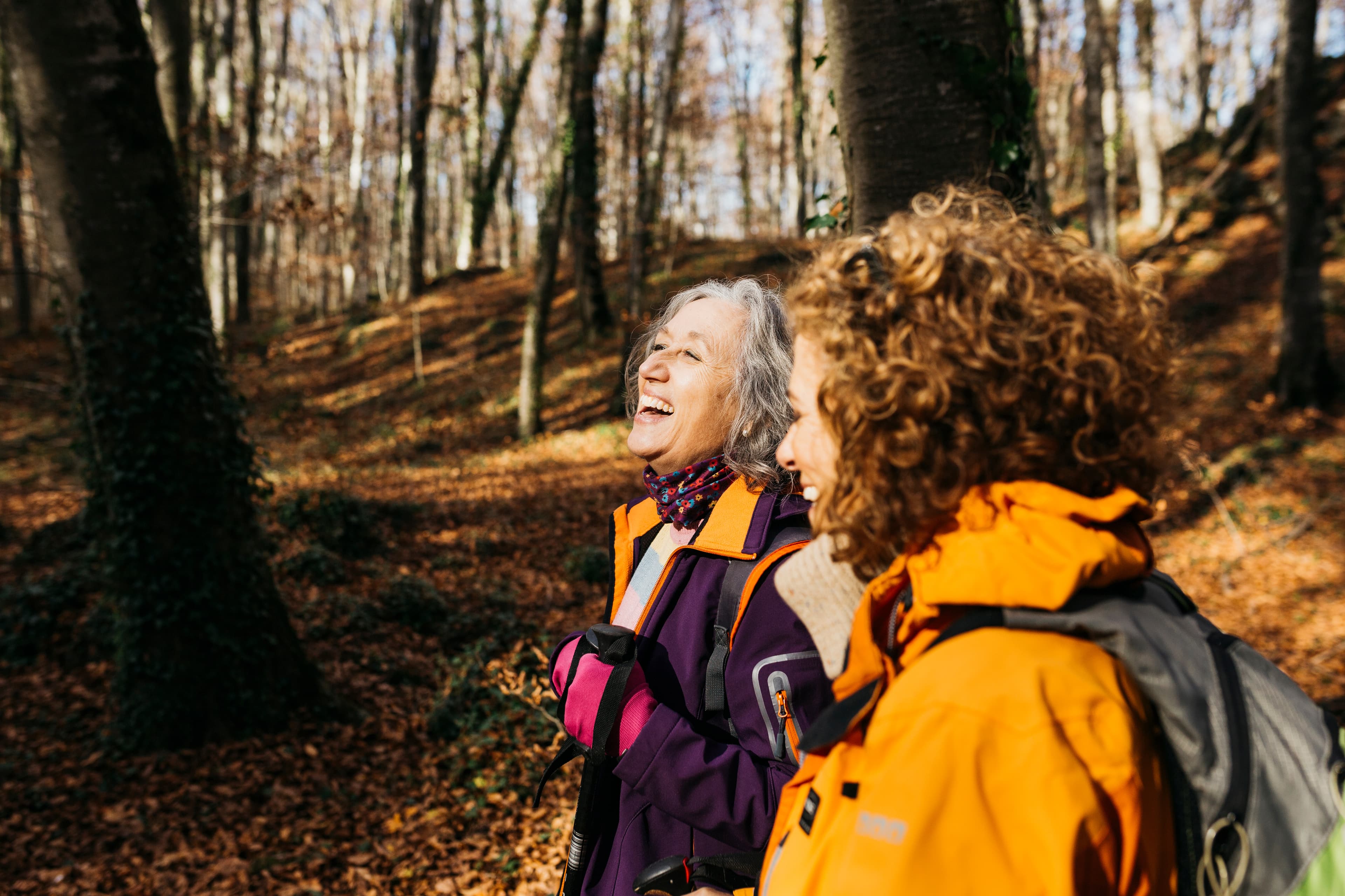 Two women in jackets laugh together while walking through a sunlit, autumn forest. - Home Instead
