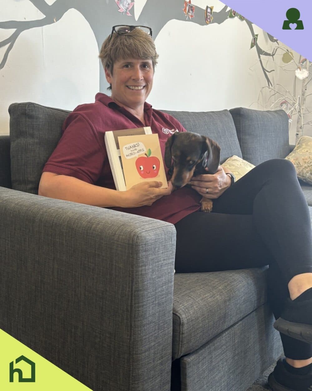Smiling person on a couch holds a book and cuddles a small dog; illustrated wall in the background. - Home Instead