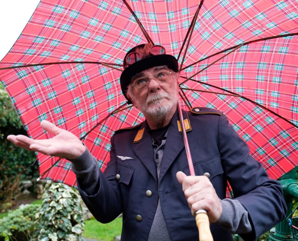 Elderly man with a tartan umbrella and wearing a uniform, brimmed hat and goggles on the hat, wondering whether it is raining.
