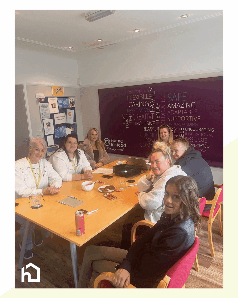 Seven people sit around a table in an office, smiling, with snacks and drinks in front of them. - Home Instead