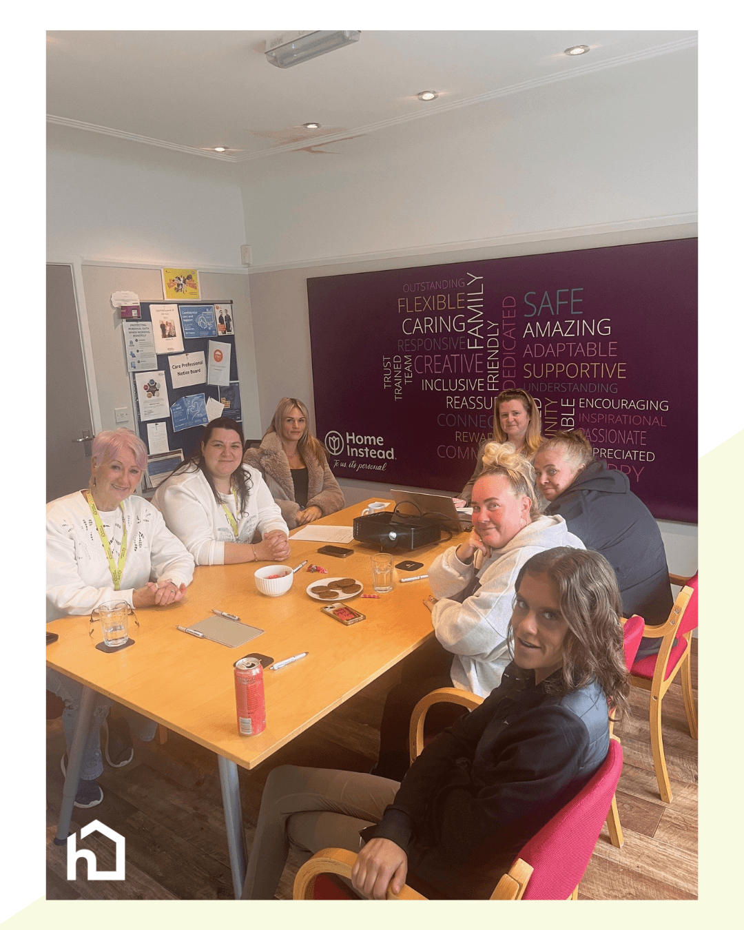 Seven people sit around a table in an office, smiling, with snacks and drinks in front of them. - Home Instead