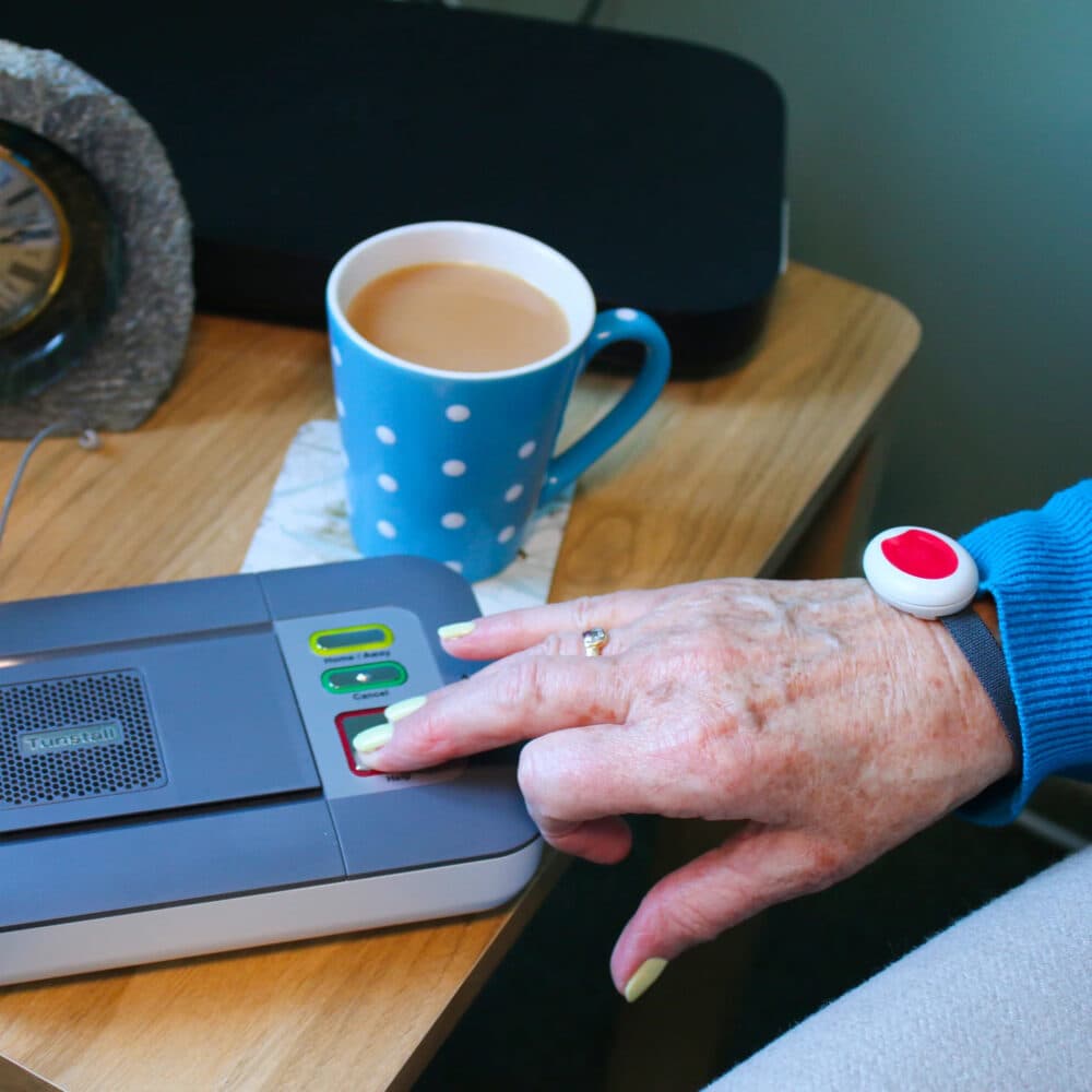 An elderly hand presses an emergency button on a device, wearing a red alert wristband, next to a cup of tea. - Home Instead