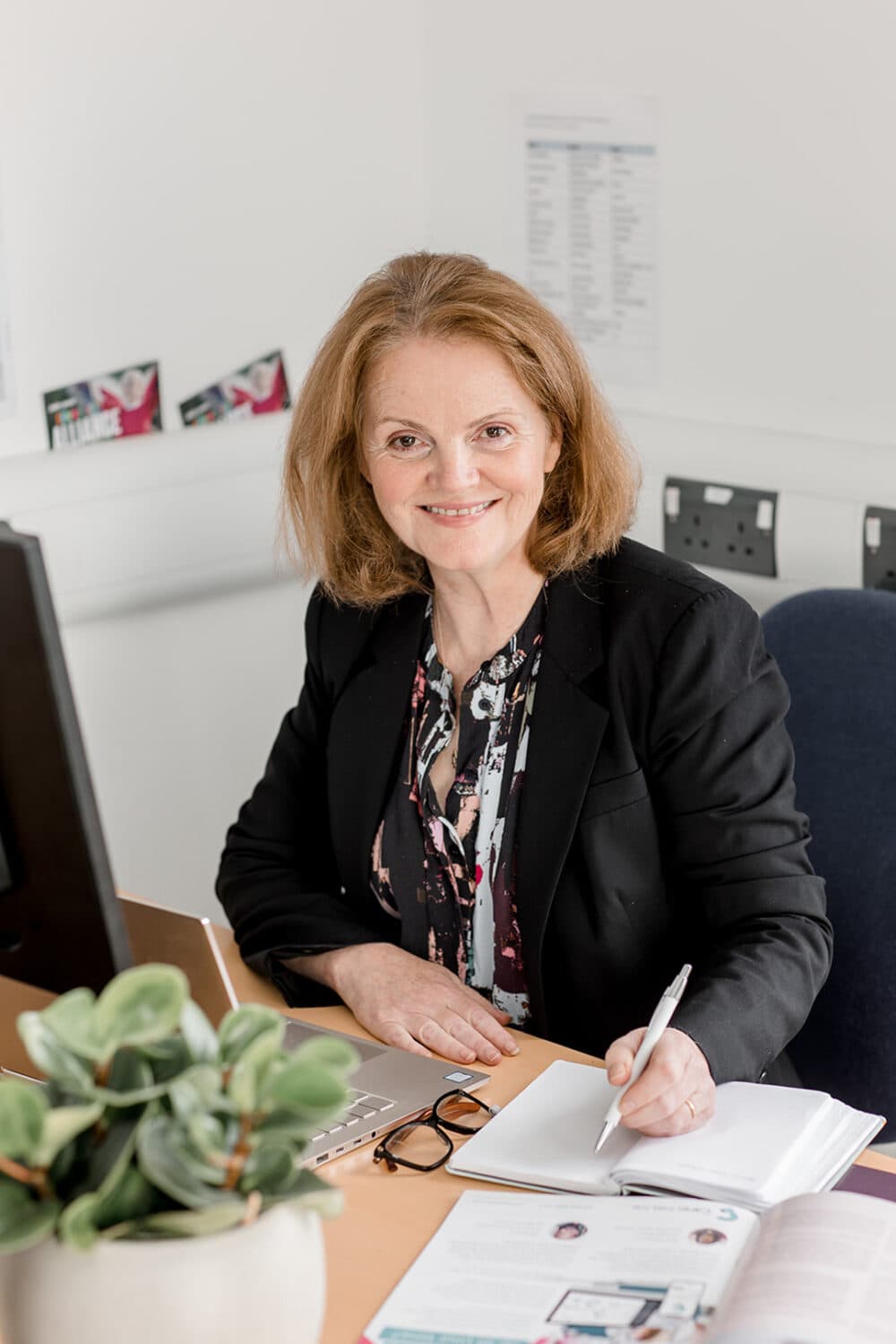 A woman in business attire sits at a desk, smiling, writing in a notebook near a computer and a plant. - Home Instead