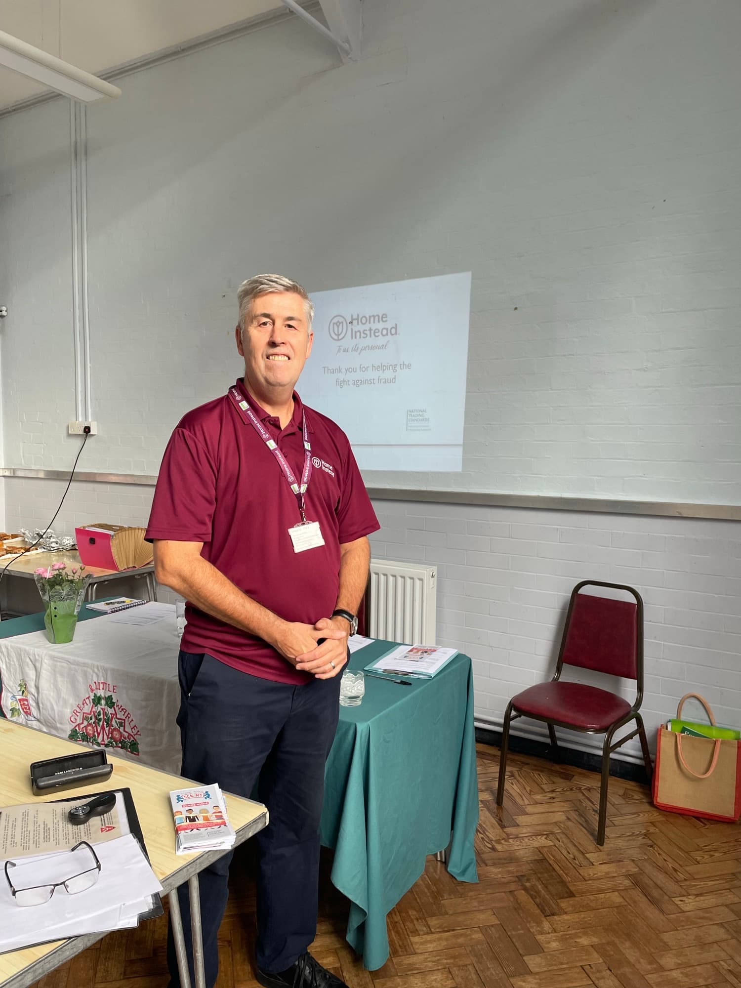A man in a maroon shirt stands by a table with brochures, in a bright room with a projected "Home Instead" slide. - Home Instead