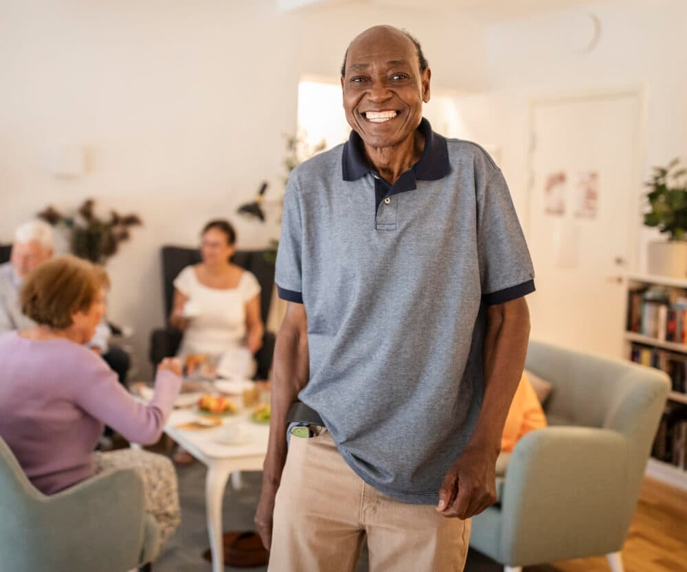 Smiling older man stands in a living room while people talk and eat at a table in the background. - Home Instead