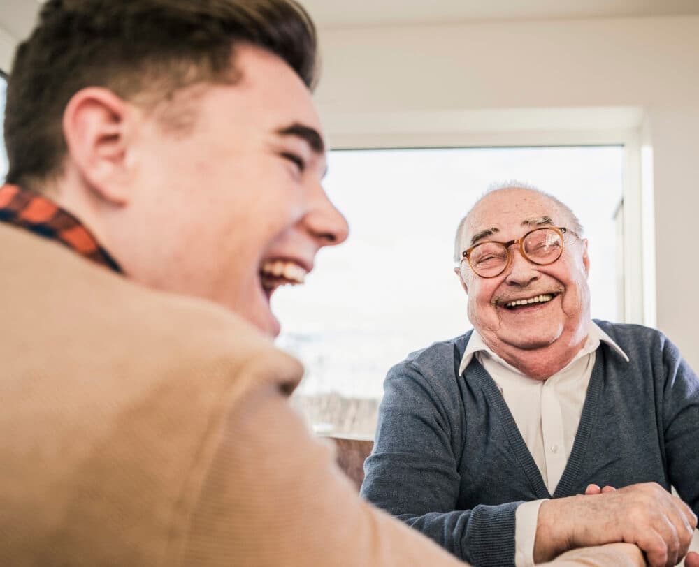 Young man and elderly man sitting together, both laughing and smiling near a bright window. - Home Instead