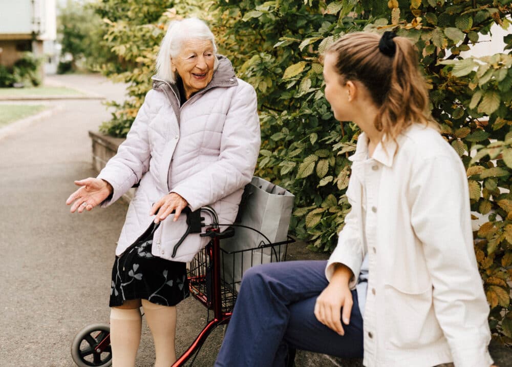 An elderly woman with a walker smiles and talks to a younger woman sitting on a low wall outdoors. - Home Instead