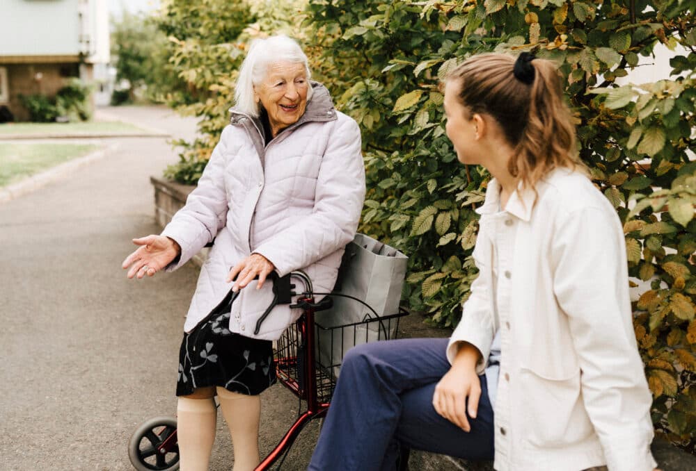 An elderly woman with a walker talks with a younger woman outdoors near green bushes. - Home Instead