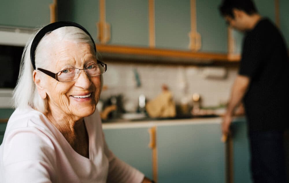 Smiling elderly woman with glasses in a kitchen, with a man in the background preparing food. - Home Instead