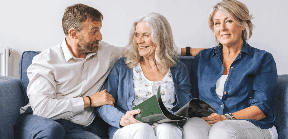 Three smiling adults sit together on a couch, holding a magazine and talking in a cozy living room. - Home Instead