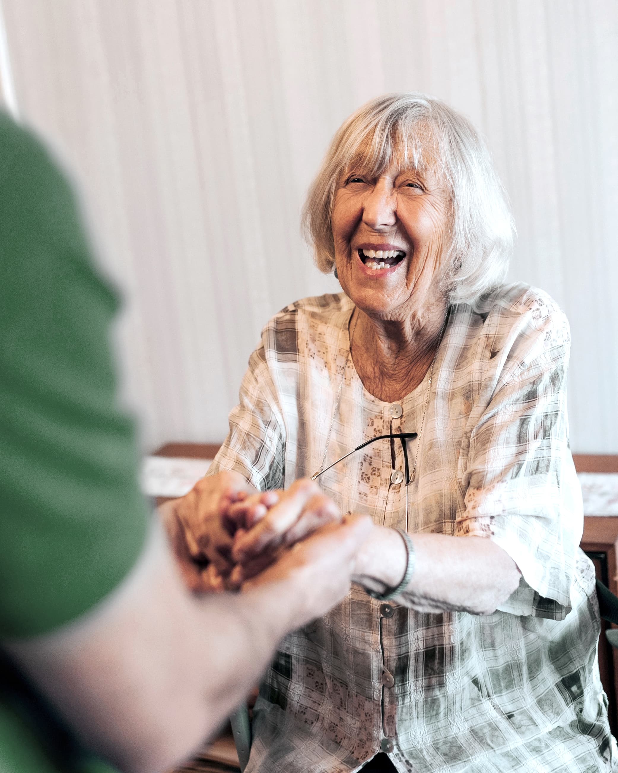 Smiling elderly woman holding hands with a person in a green shirt, sitting indoors. - Home Instead