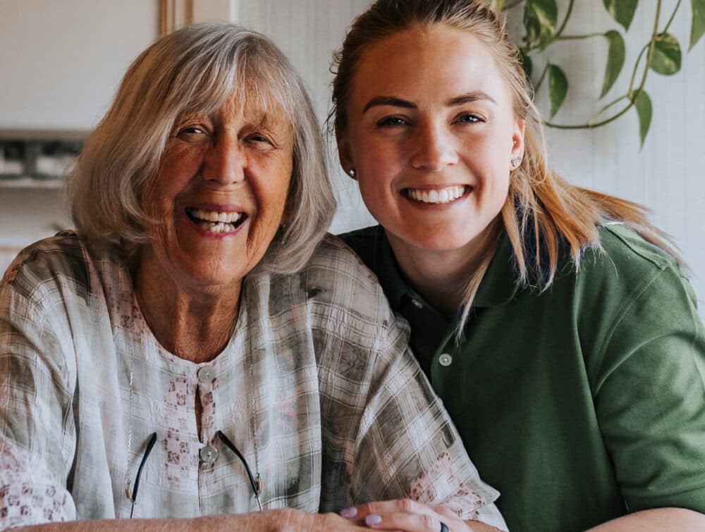 Older woman and younger woman smiling together indoors, sitting closely at a table. - Home Instead