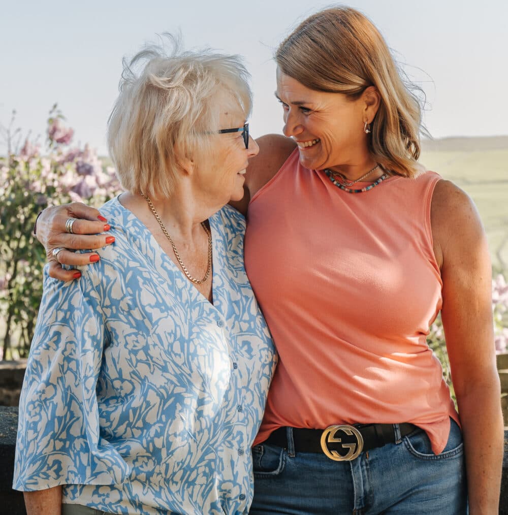 Two women smiling and embracing each other outdoors, standing in front of blooming flowers. - Home Instead
