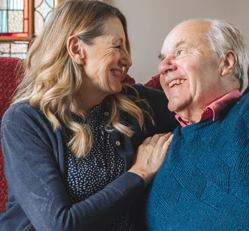 Smiling woman and elderly man sit close together, sharing a happy moment on a red couch. - Home Instead