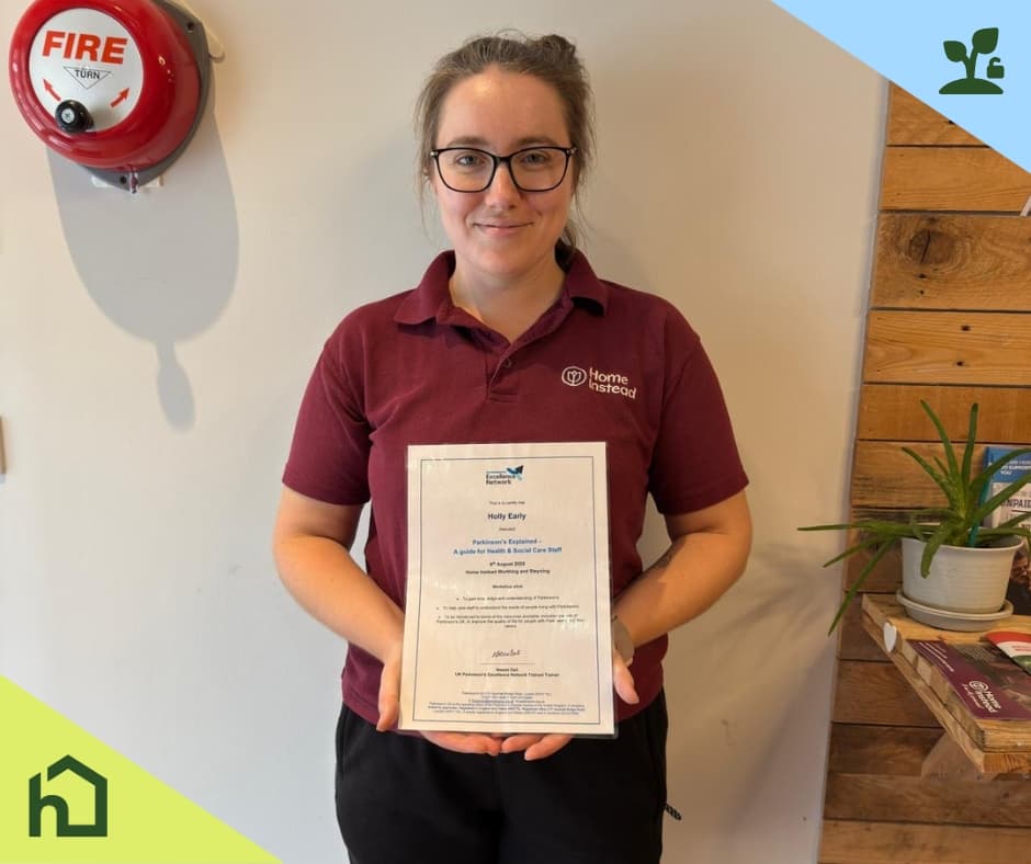 A woman in a maroon shirt holds a certificate and smiles, standing indoors by a fire alarm and a plant. - Home Instead