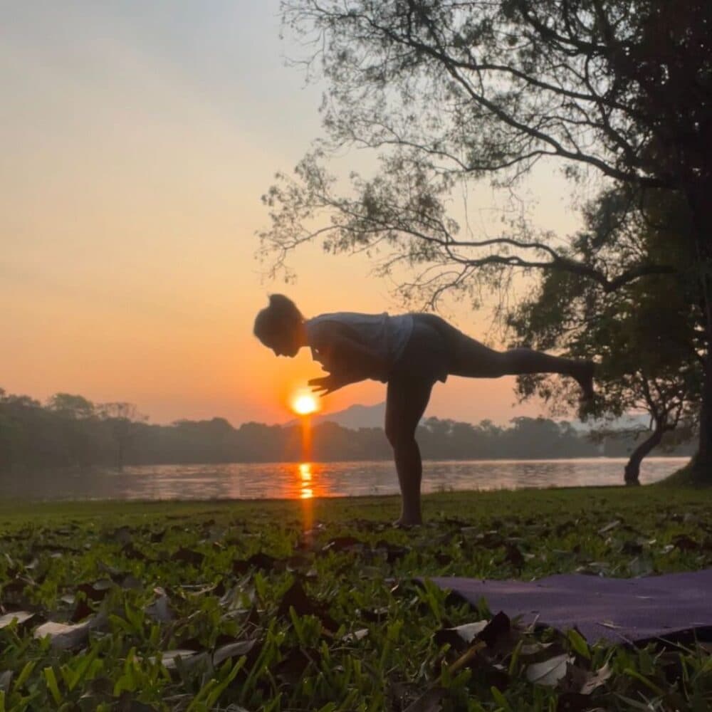 Person doing a yoga pose outdoors at sunrise by a lake, with trees and grass in the background. - Home Instead