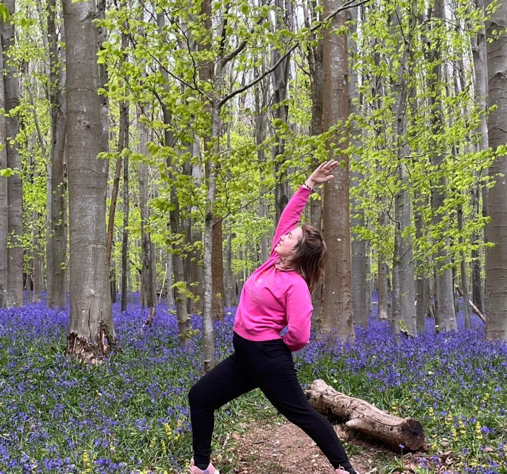 Person in a pink hoodie doing a yoga pose in a forest with bluebells and tall trees. - Home Instead