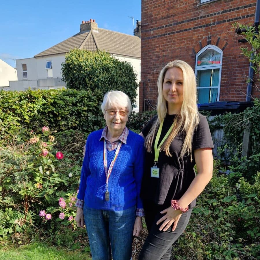 Two women, one older and one younger, standing together in a garden with flowers and greenery. - Home Instead