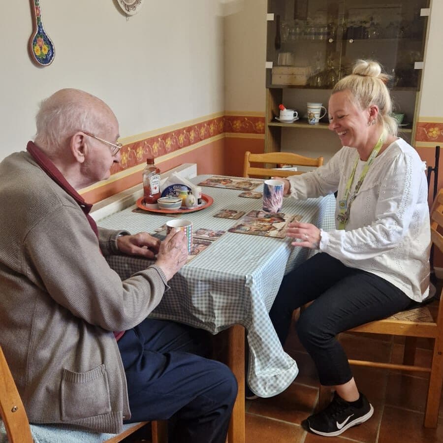 An elderly man and a woman sit at a kitchen table, smiling and drinking from mugs. - Home Instead