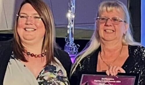 Two women smiling and holding an award certificate at an indoor event. - Home Instead