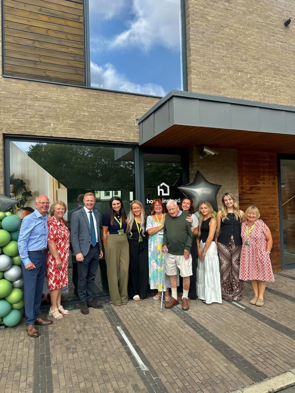 A group of people smiling and posing together outside a building with balloons and a large star decoration. - Home Instead