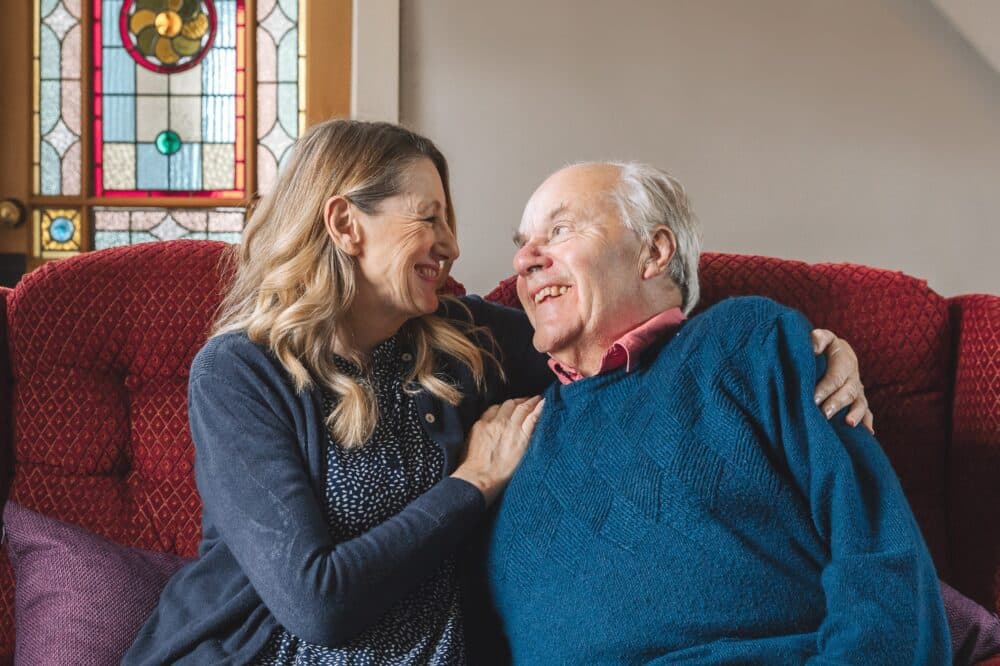 Elderly man sitting on sofa with a younger woman, looking at each other and smiling