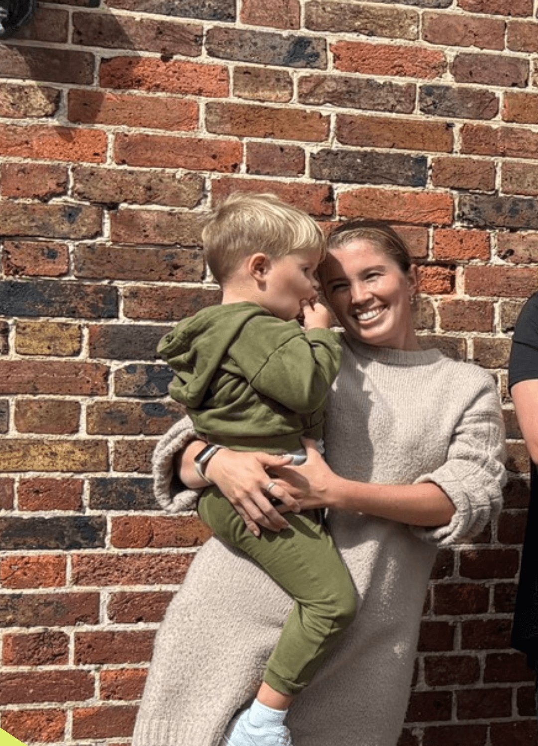 A smiling woman in a beige sweater holds a young boy dressed in a green hoodie and pants against a brick wall.
