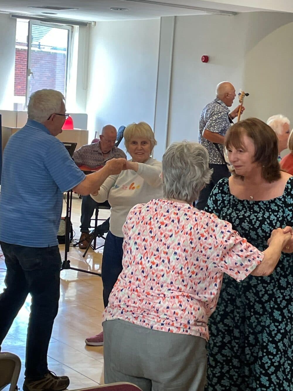 Seniors dance together in a bright room, smiling and holding hands in pairs. - Home Instead