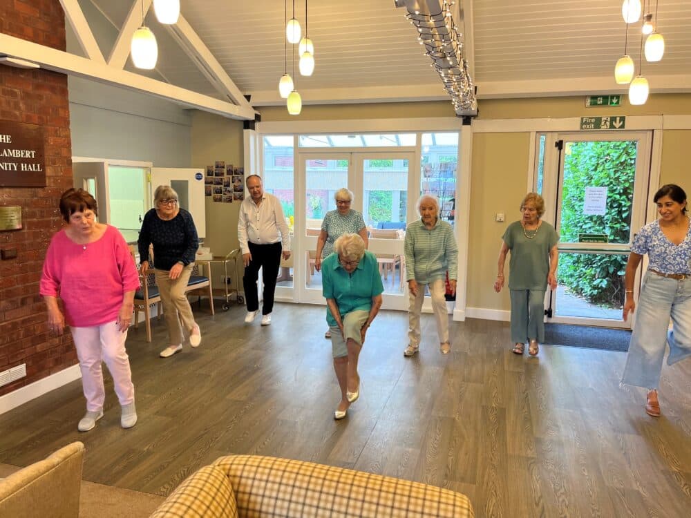 A group of seniors and a young woman participate in a dance or exercise class in a bright community room. - Home Instead
