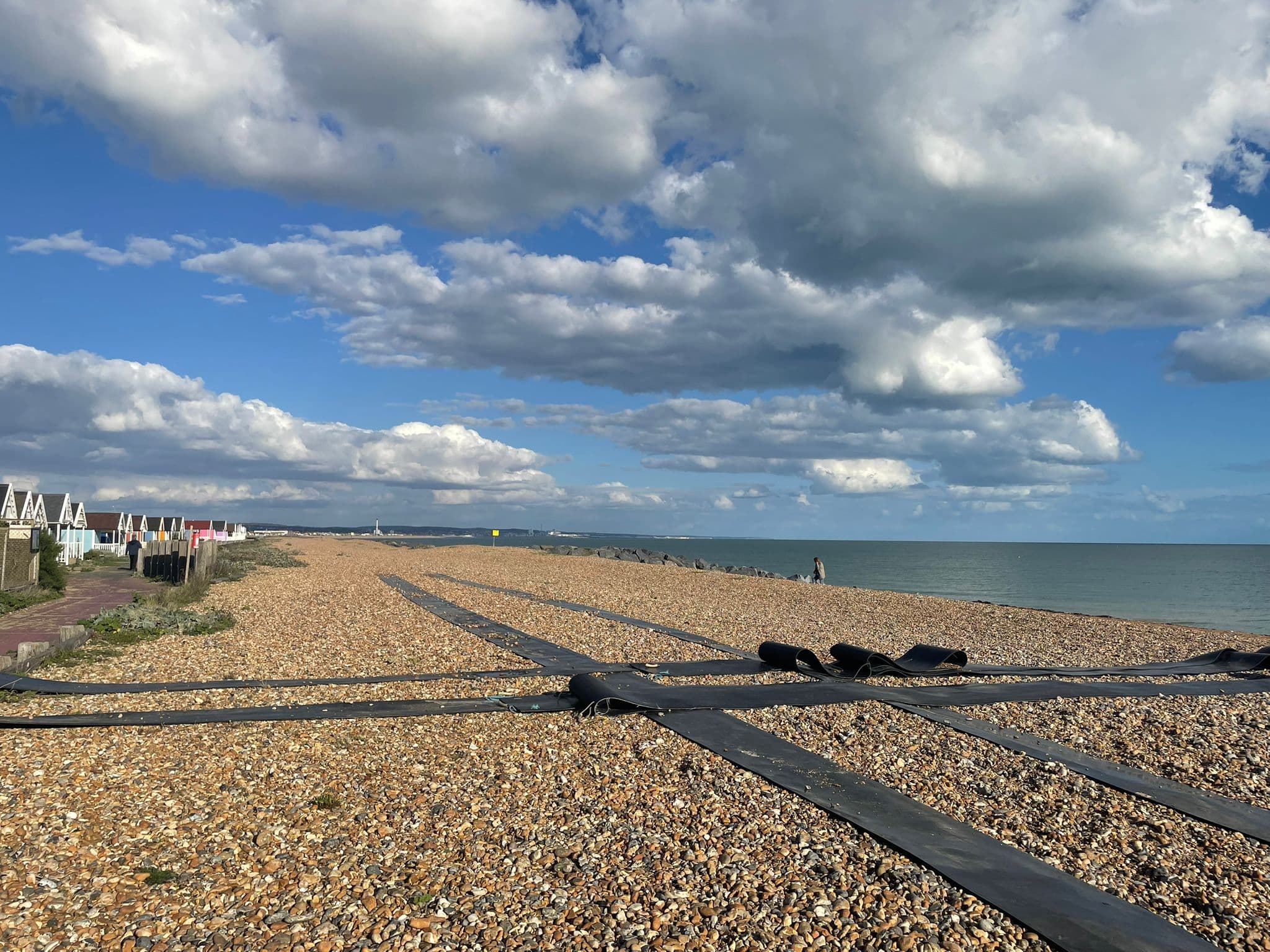 Pebble beach with black pipes, blue sky, white clouds, and sea; beach huts on the left. - Home Instead