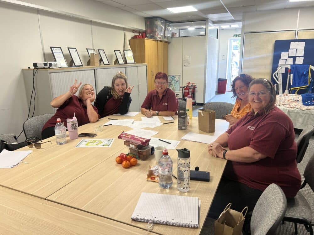 Five women sit around a table smiling and working with papers in a bright office meeting room. - Home Instead