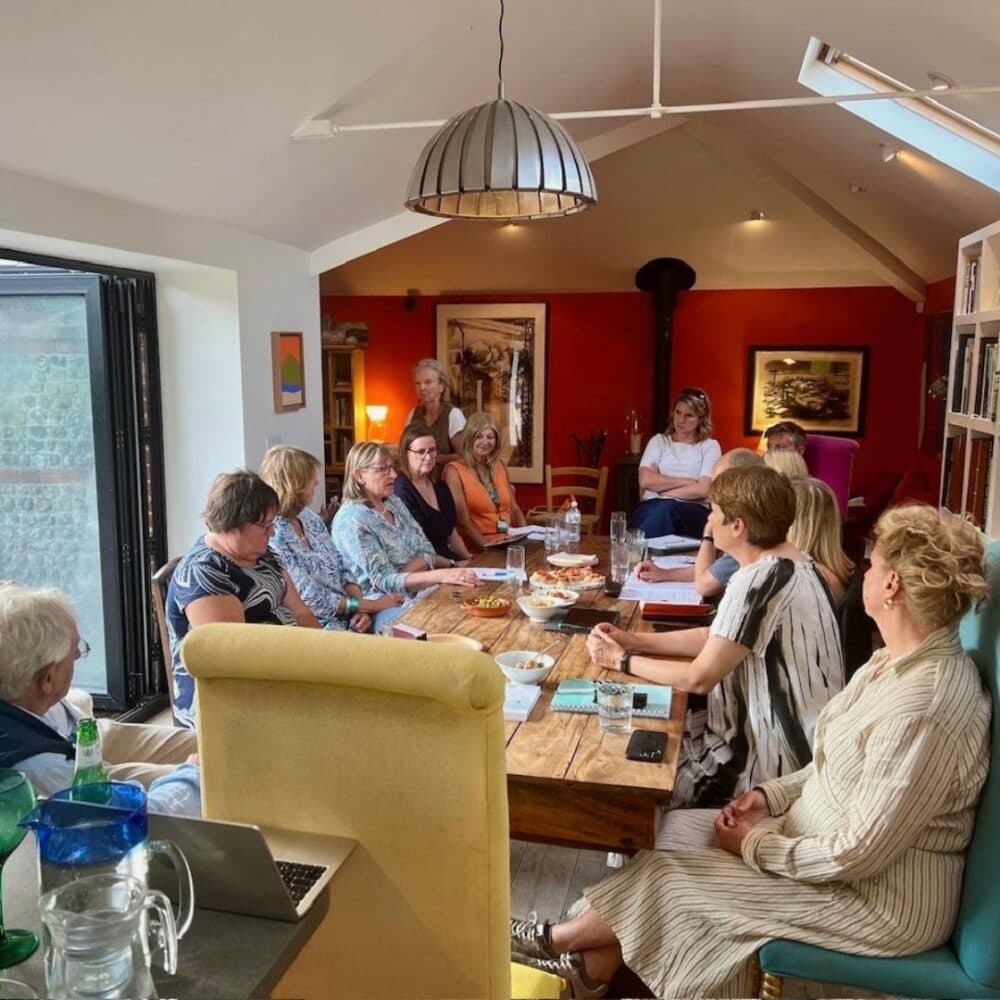 A group of people sit around a long wooden table having a meeting in a cozy, well-lit room. - Home Instead