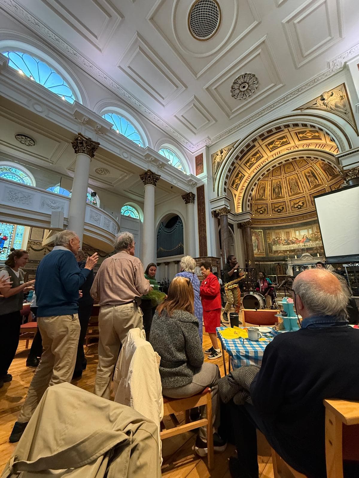 People gathered in a grand hall with ornate ceiling, arches, and stained glass windows, some standing and some seated. - Home Instead
