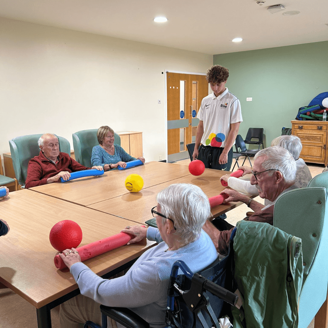 Six seniors play with balls and foam sticks at a table, guided by a young instructor in a care home setting. - Home Instead
