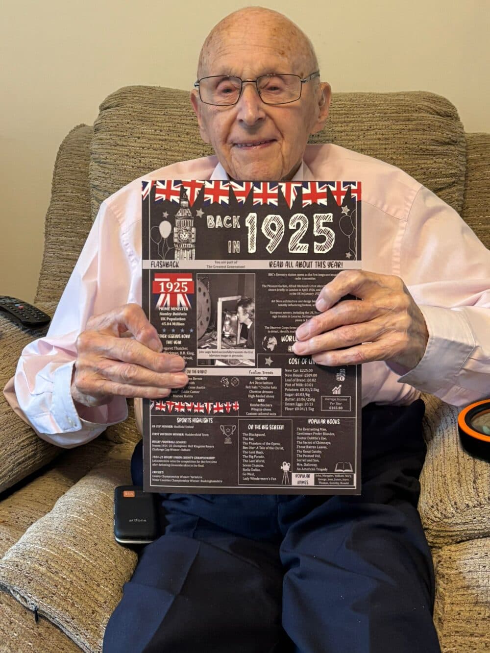 Smiling elderly man on a sofa holds a “Back in 1925” sign with historical facts and British flags. - Home Instead