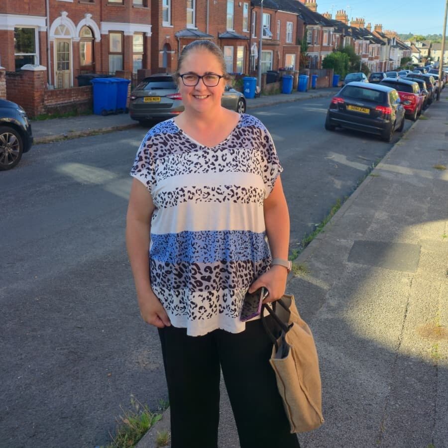 Woman stands on a sunny street, smiling, holding a tan bag, with houses and parked cars in the background. - Home Instead