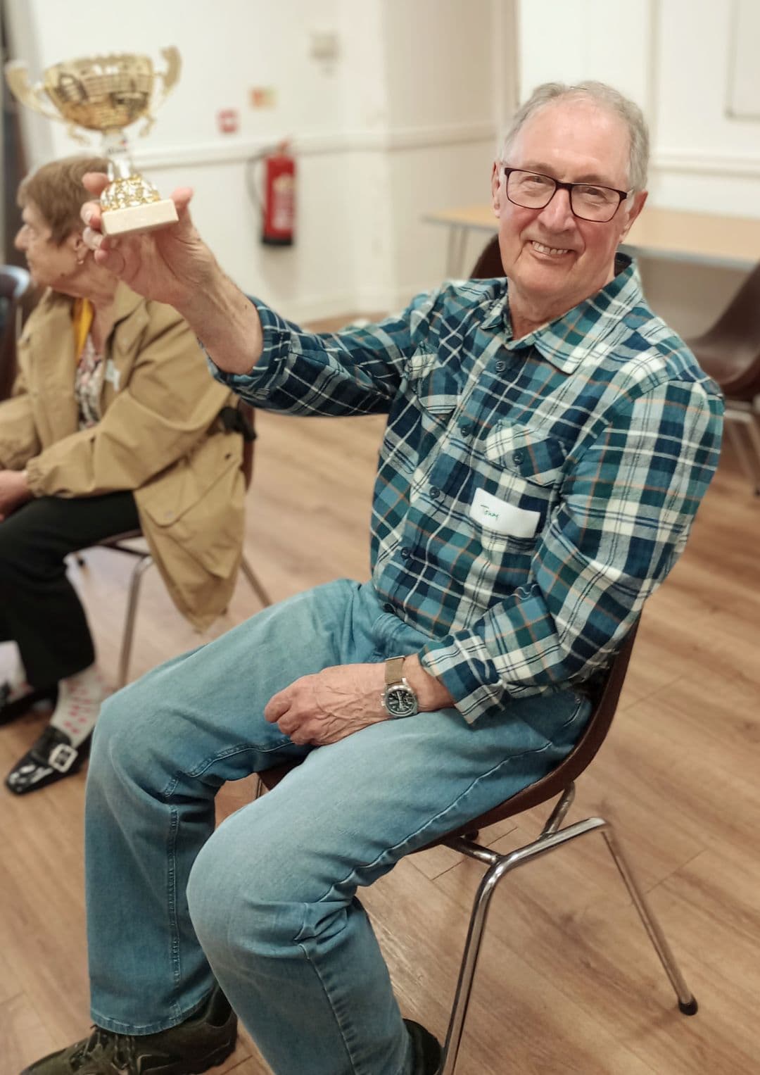 Smiling man in glasses holds up a trophy while sitting on a chair indoors; another person sits beside him. - Home Instead