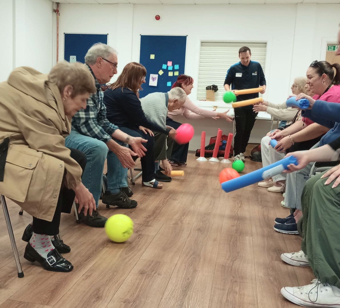 A group of people sitting in two rows play an indoor ball game using plastic sticks and colorful balls. - Home Instead
