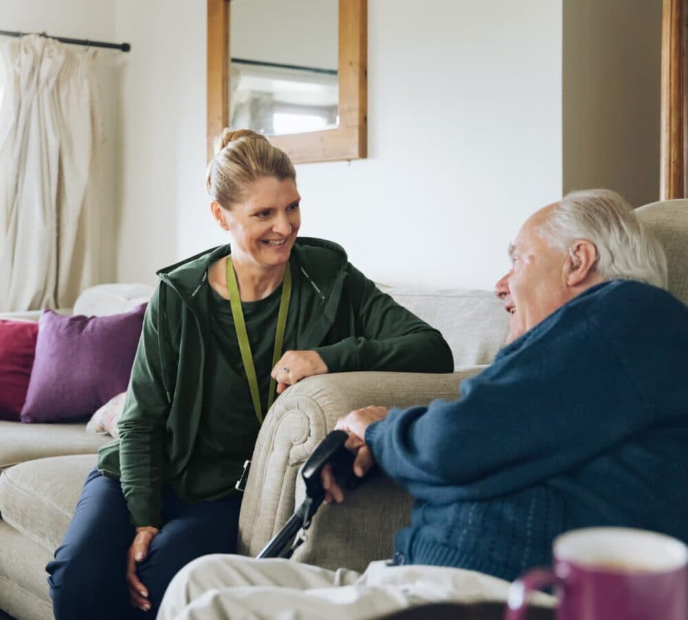 Two people happily chatting together while sitting on a couch inside the house Home Instead