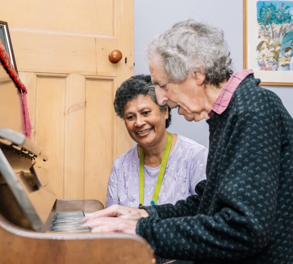 An older male adult with grey hair and wearing dark sweater playing the piano while his younger female carer with short hair is happily watching her